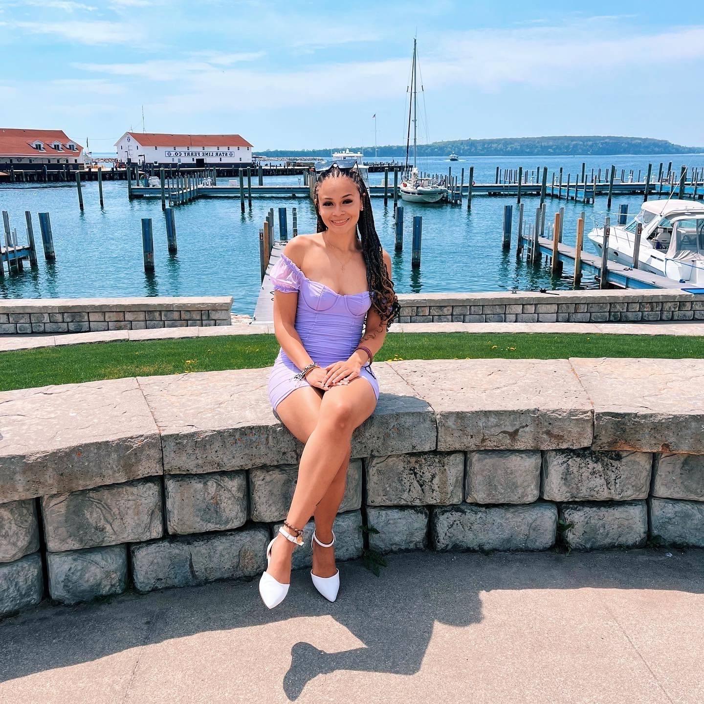 Girl with black hair and purple dress sitting on a stone wall with a lake and harbor in the background